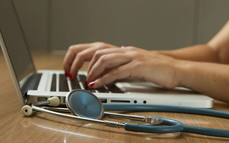 Hände tippen auf einem Laptop, daneben liegt ein Stethoskop