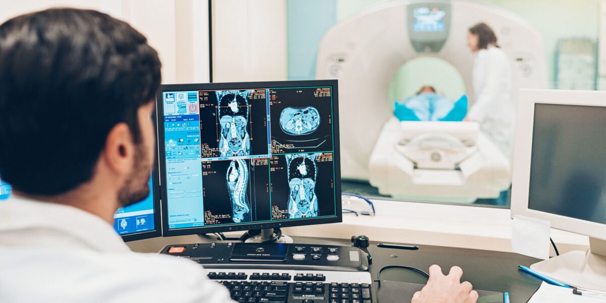 Apparategemeinschaft: Mann betrachtet den Scan eines Patienten am Computer. Im Hintergrund  sieht man eine Frau und den Patienten.